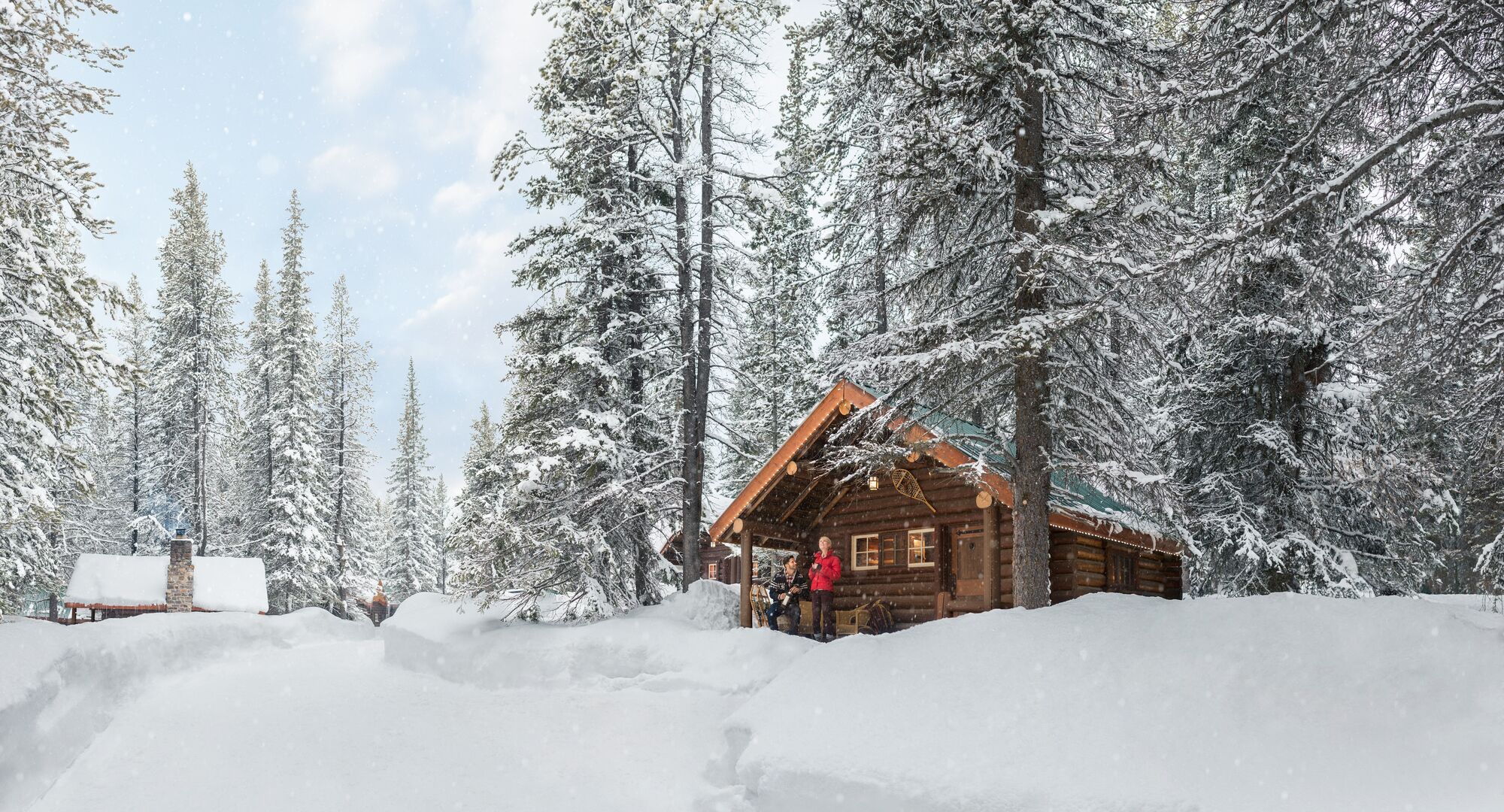 A cabin in Banff National Park covered in snow with two people on the balcony taking in the winter scenery.