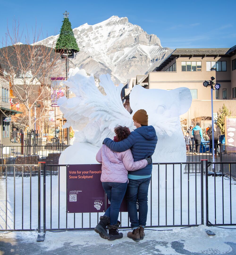 Two people standing in front of a giant snow sculpture during the SnowDays festival in Banff National Park