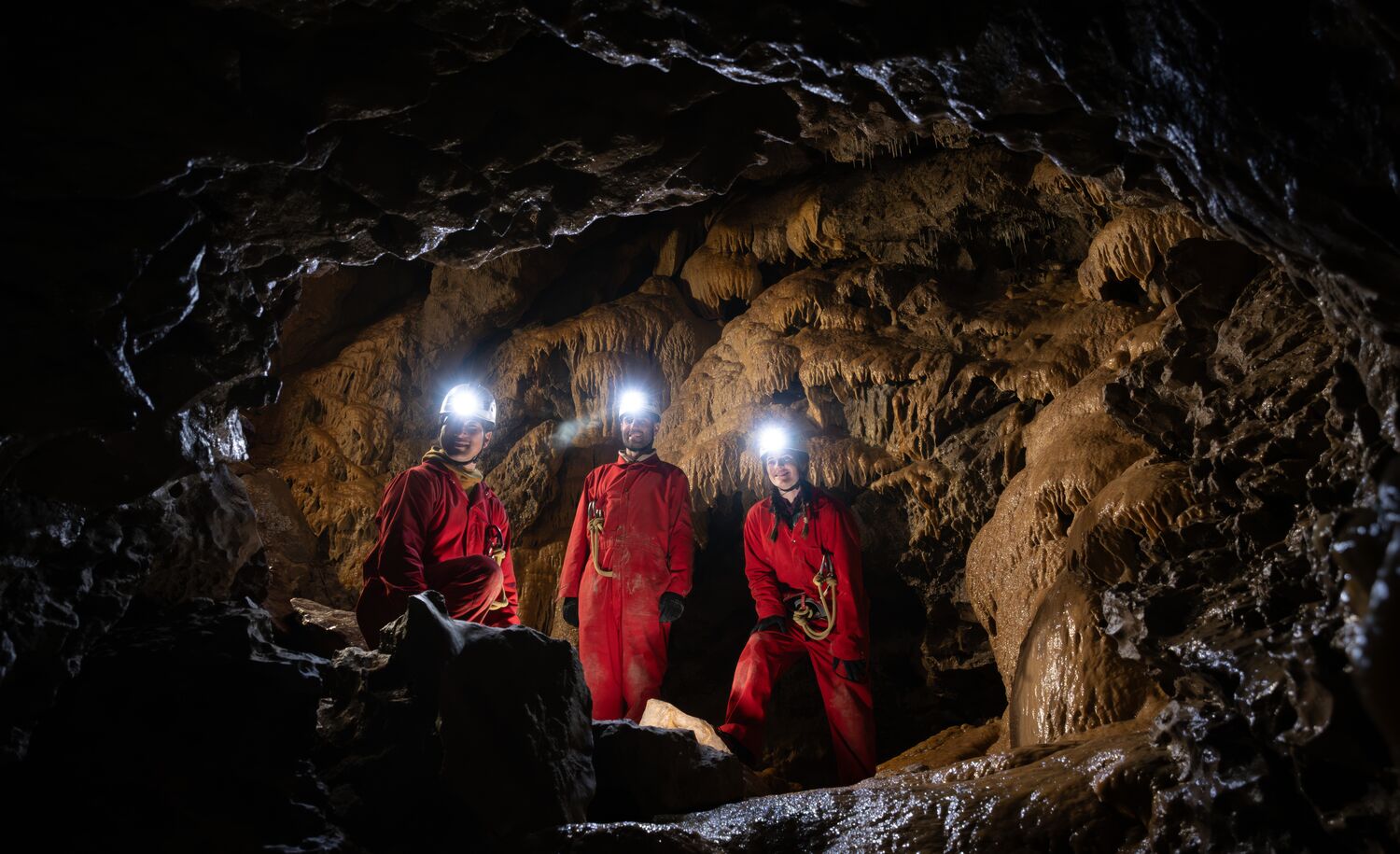 A group moves through a dark underground cave wearing protective gear and headlamps while exploring with Canmore Cave Tours.