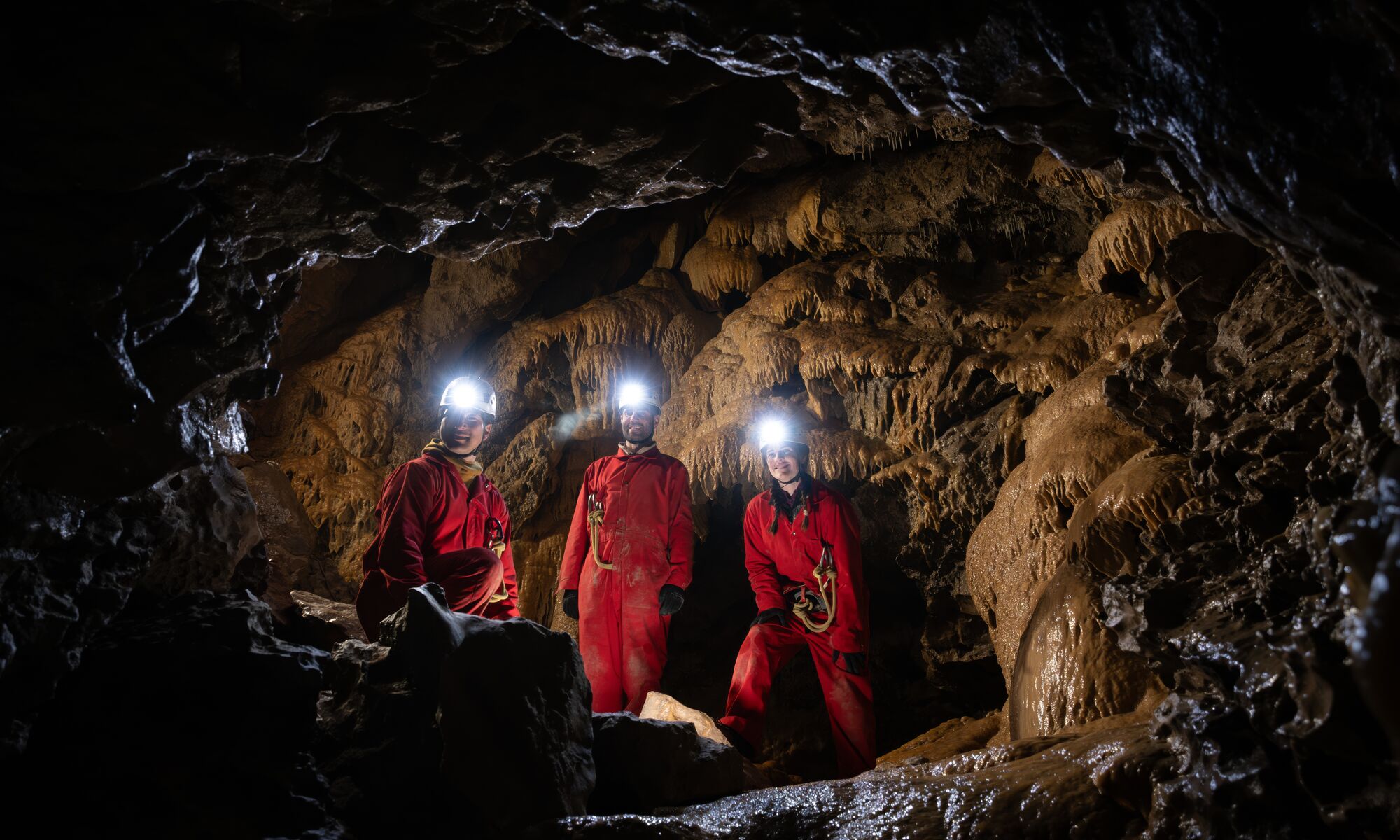 A group moves through a dark underground cave wearing protective gear and headlamps while exploring with Canmore Cave Tours.