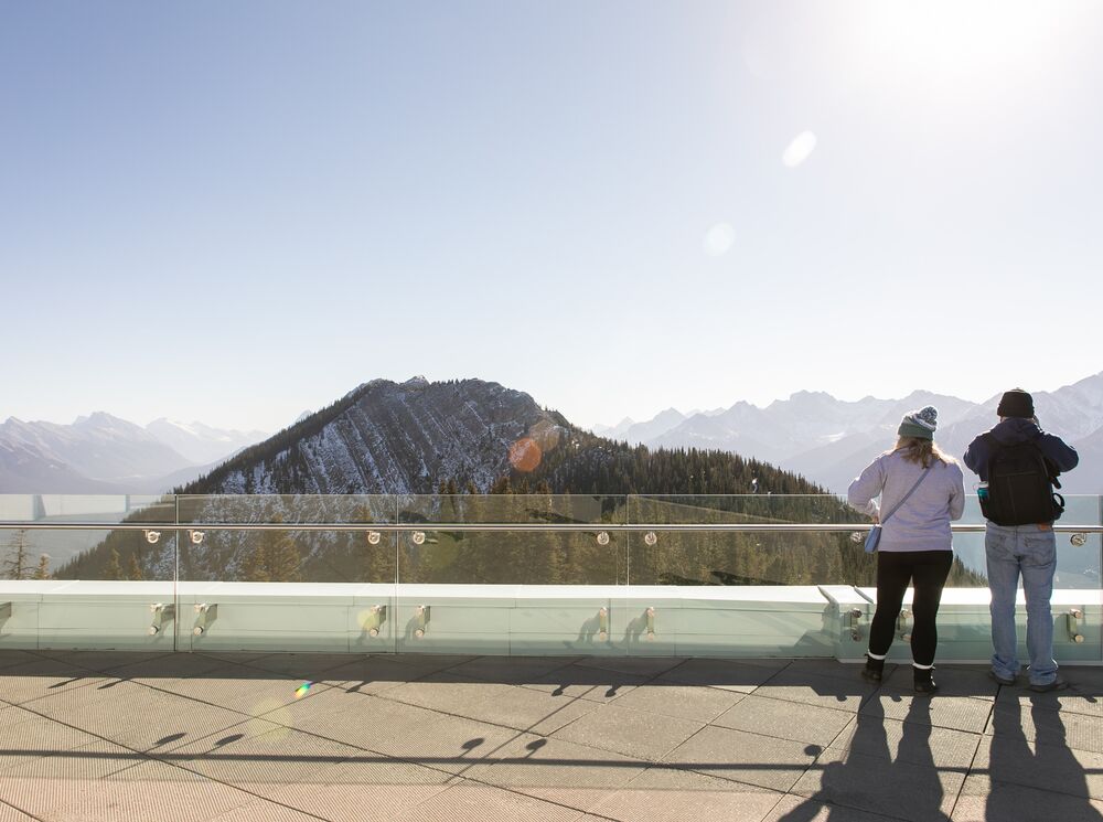 Two people look over the edge at the Banff Gondola with a mountain in the background.