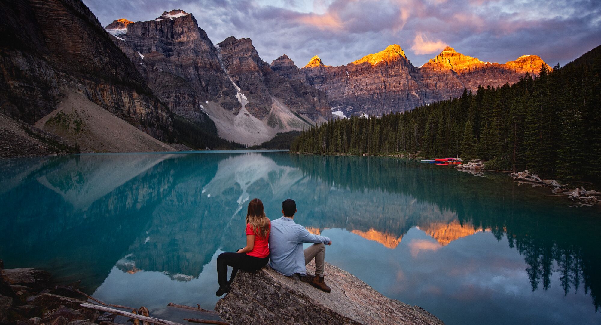 Two people sit on a rock by the water at Moraine Lake at sunrise in the summer. Alpen glow hits the tips of the mountain peaks and reflects in the water.