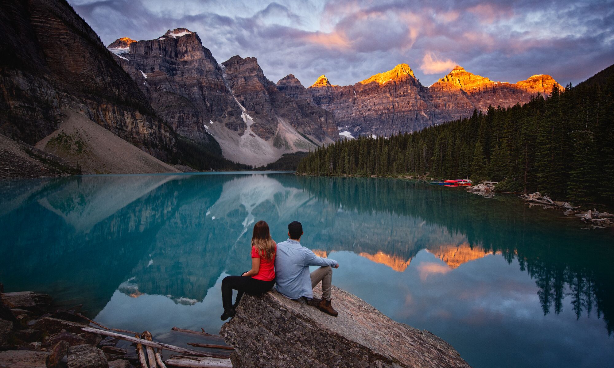 Two people sit on a rock at Moraine Lake at sunrise.