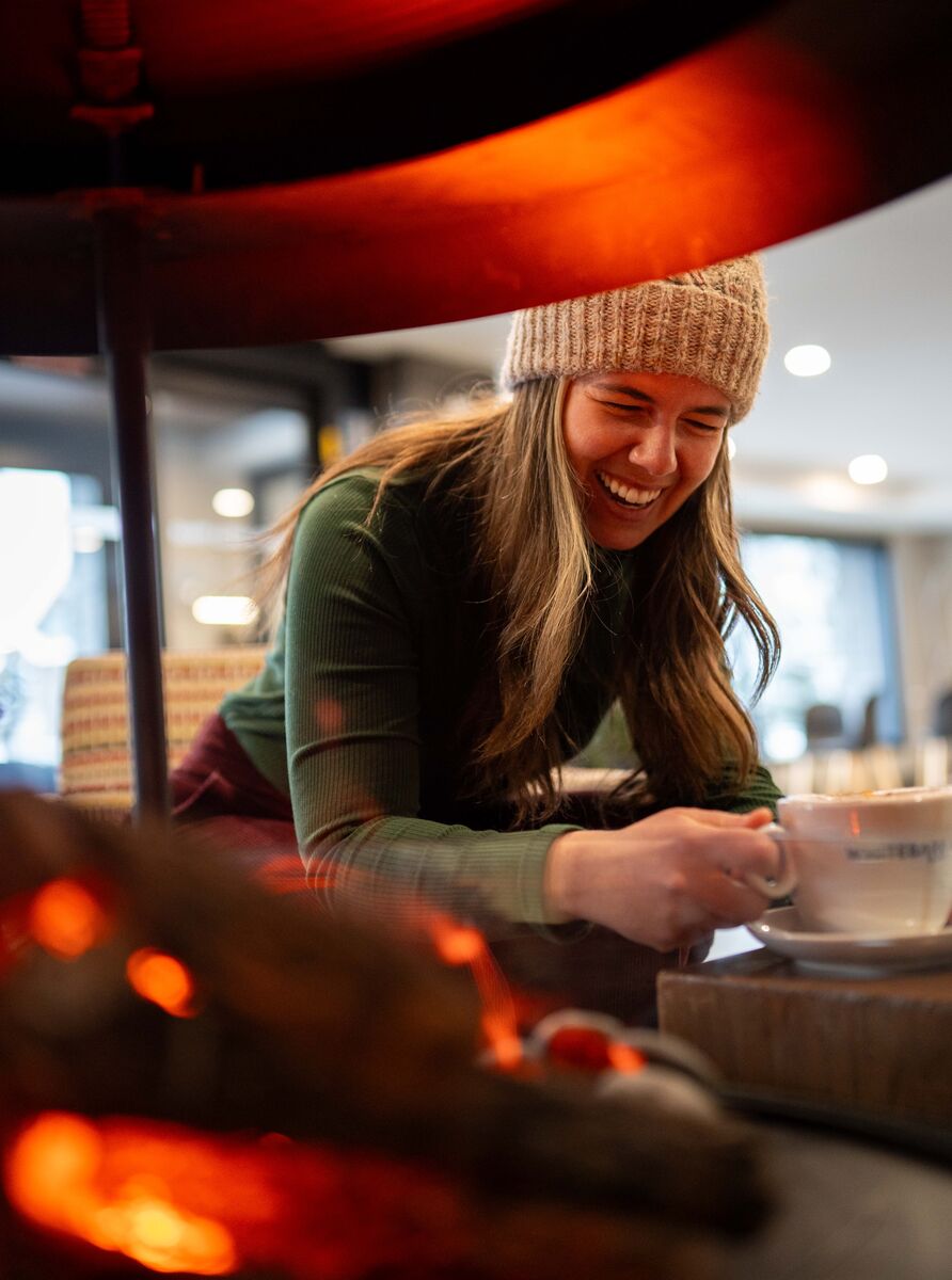 A woman laughs while drinking a hot chocolate near a fire pit.