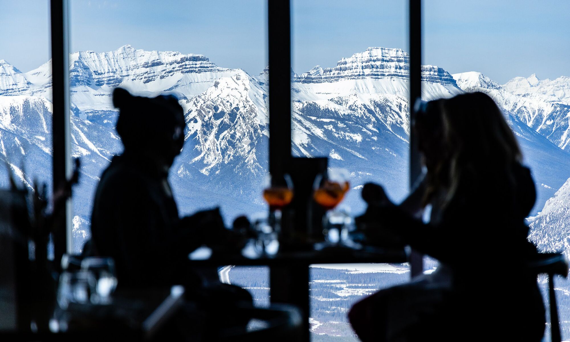 Four people dine with the Sundance Mountain Range behind them out of a window at the Banff Gondola in Banff National Park.