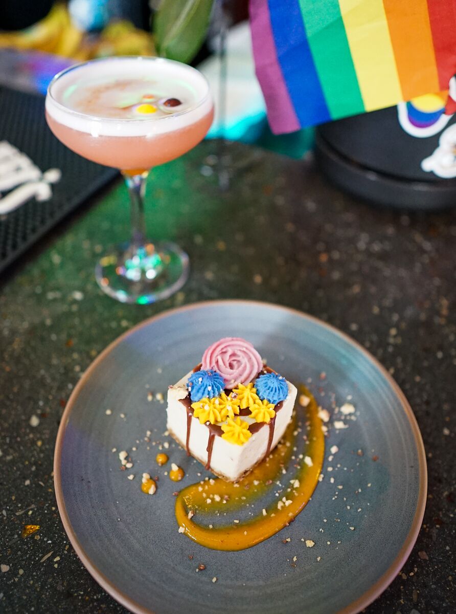 A colourful cake desert on a plate with a pink drink beside it and a pride flag at Nourish Bistro in Banff National Park. 