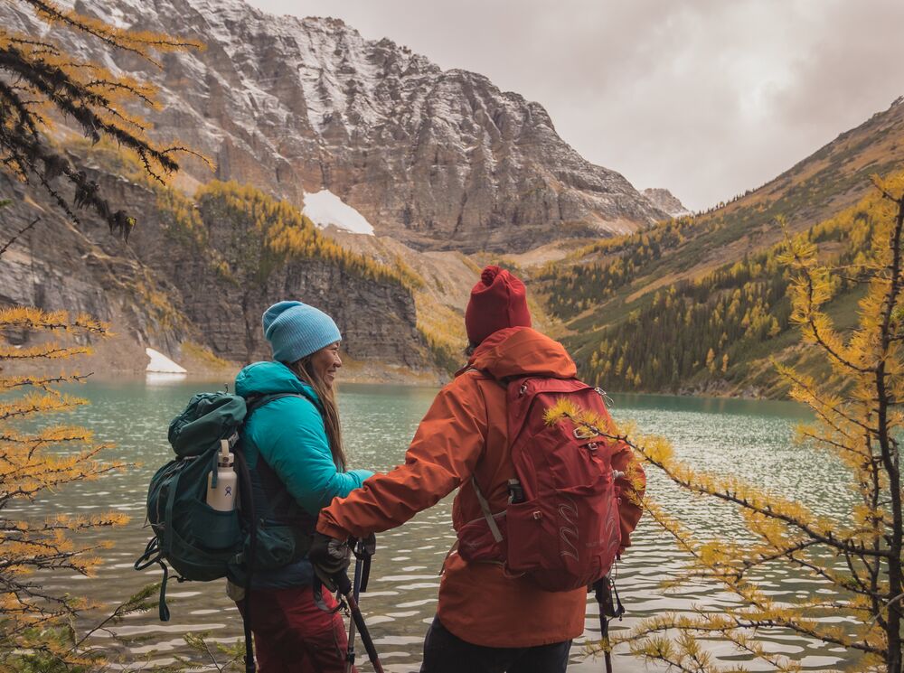 A man and woman hike alongside a lake with yellow larch trees and fresh snow on the mountains surrounding them