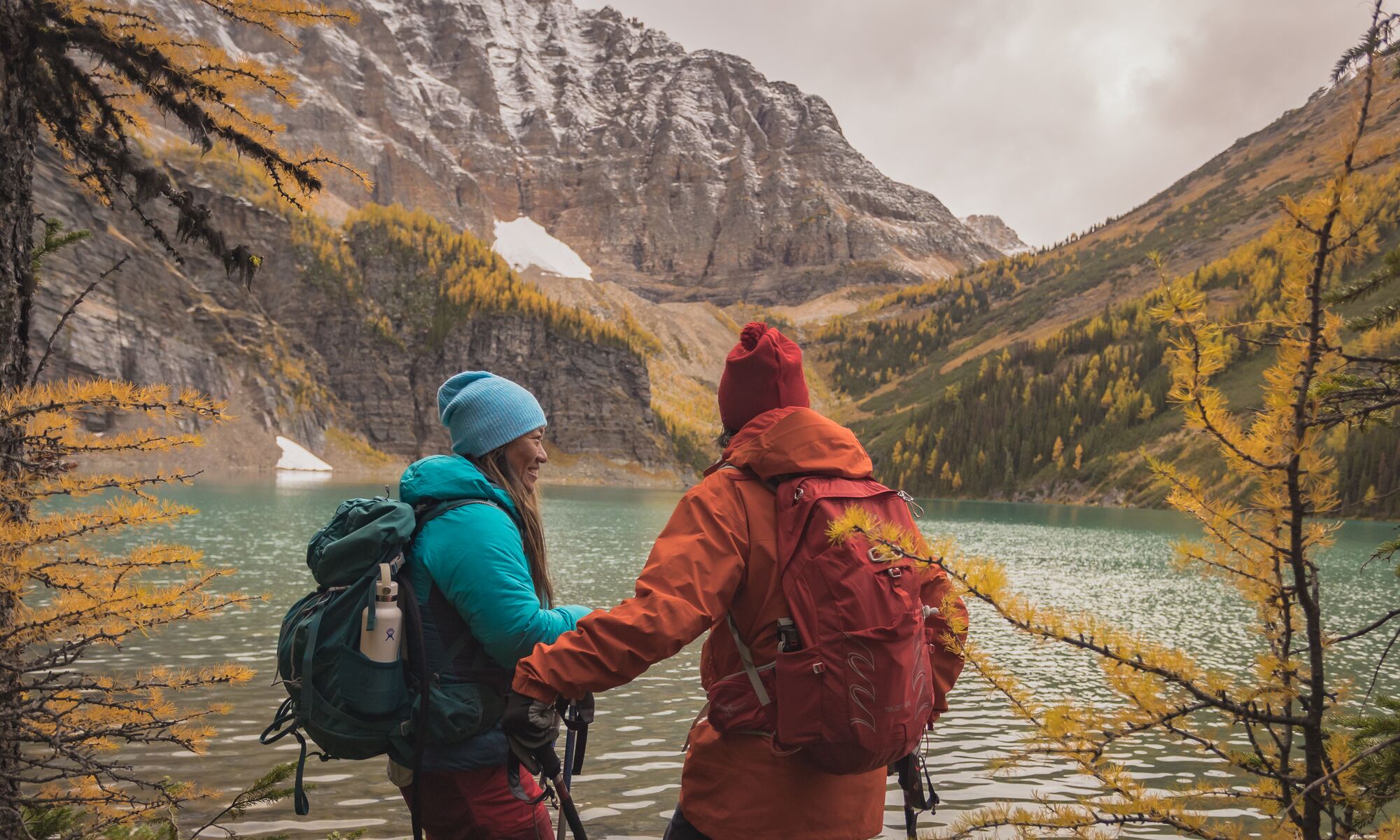 A man and woman hike alongside a lake with yellow larch trees and fresh snow on the mountains surrounding them