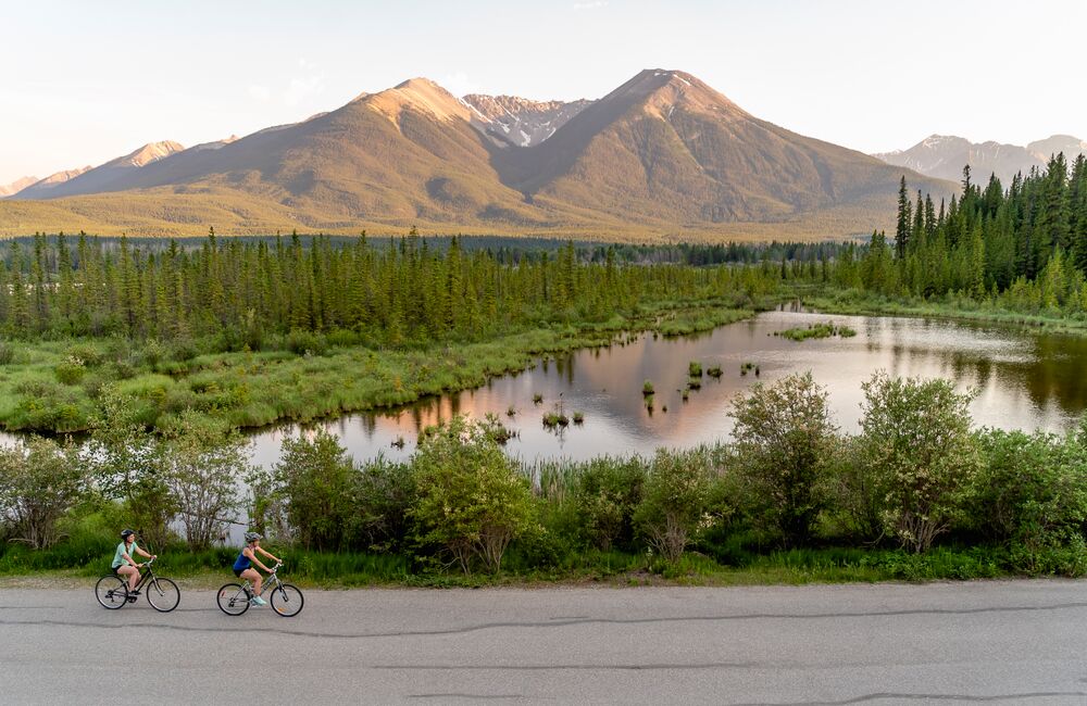 A couple of friends biking along the Vermilion Lakes in Banff National Park with a lake and a mountain behind them at sunset.