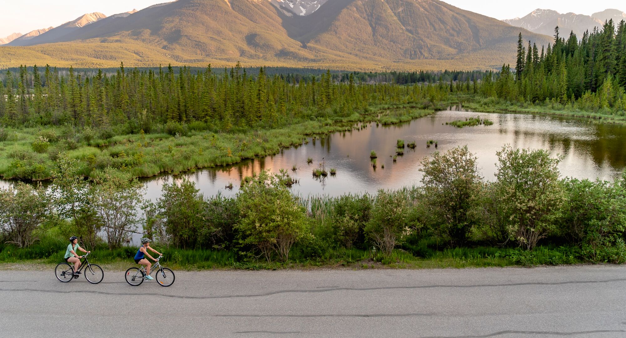 A couple of friends biking along the Vermilion Lakes in Banff National Park with a lake and a mountain behind them at sunset.