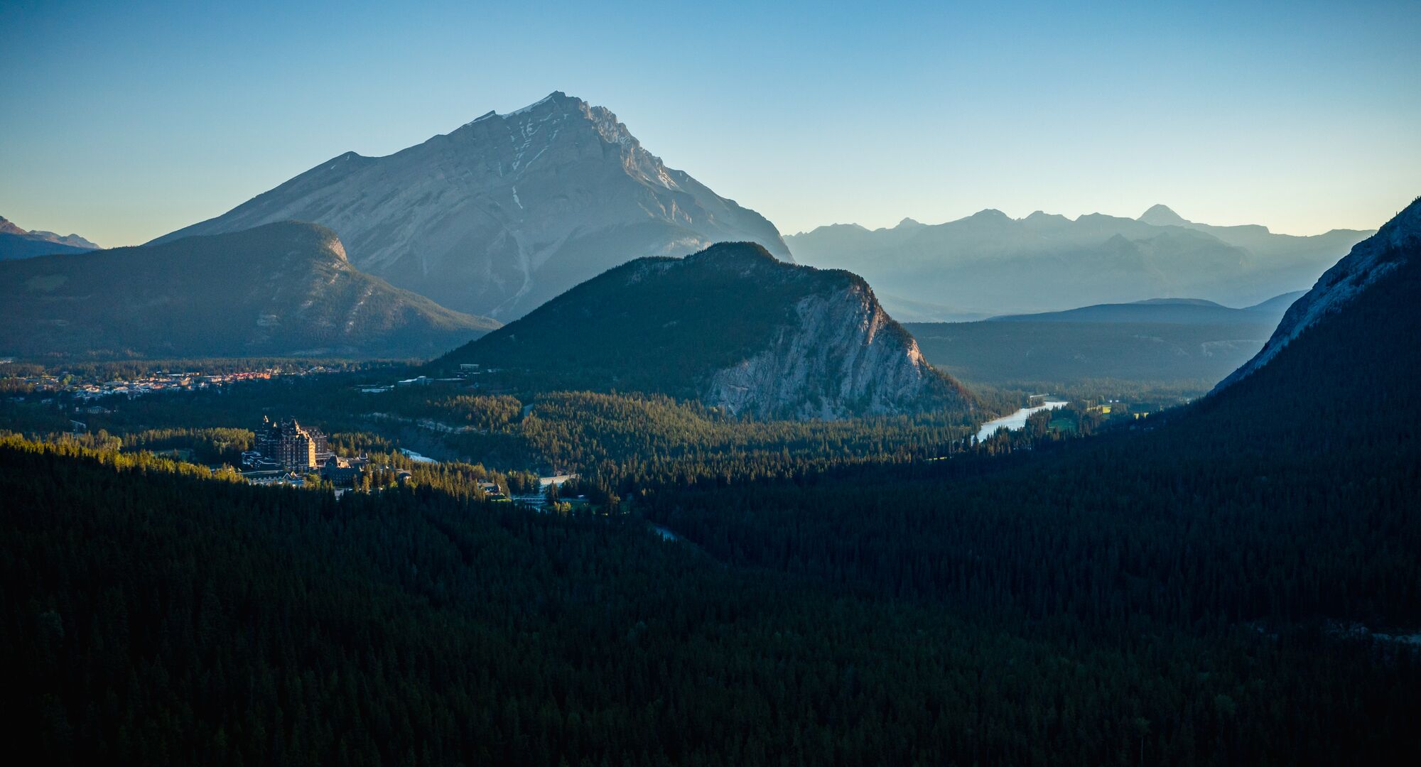 Wide view of forested valleys and layered mountains at sunrise, with soft light touching the peaks and a river winding through the landscape.