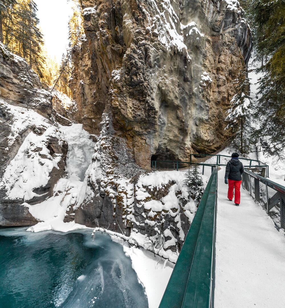 A woman walks across a bridge beside a frozen waterfall while hiking Johnston Canyon in the spring in Banff National Park
