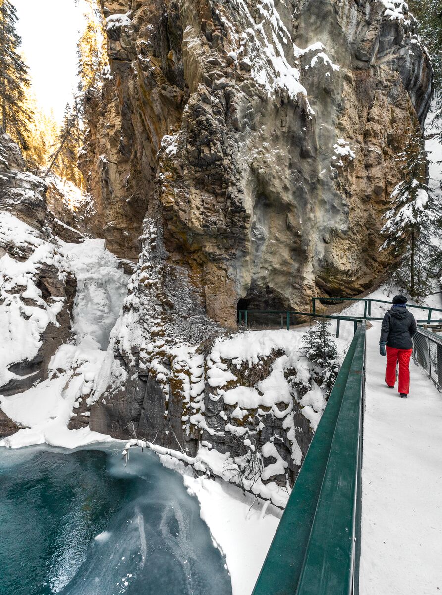 A person walks on a bridge with a frozen waterfall behind it at Johnston Canyon in winter in Banff National Park.