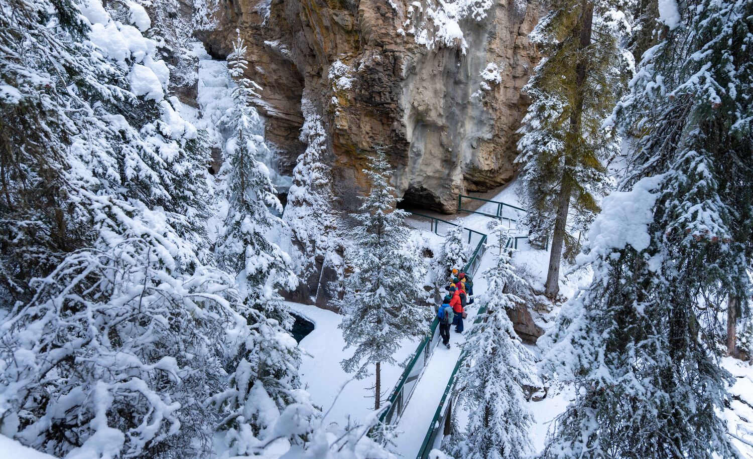 A tour group stands on a snowy bridge at Johnston Canyon Lower Falls as seen from above in Banff National Park.