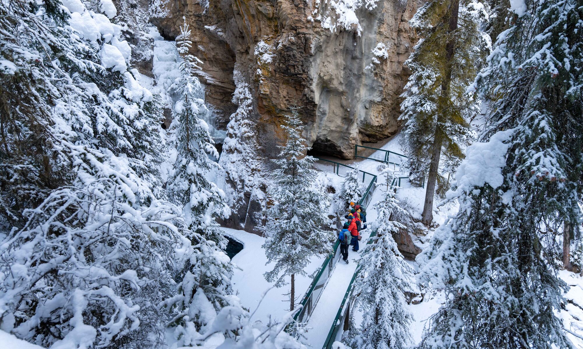 A tour group stands on a snowy bridge at Johnston Canyon Lower Falls as seen from above in Banff National Park.