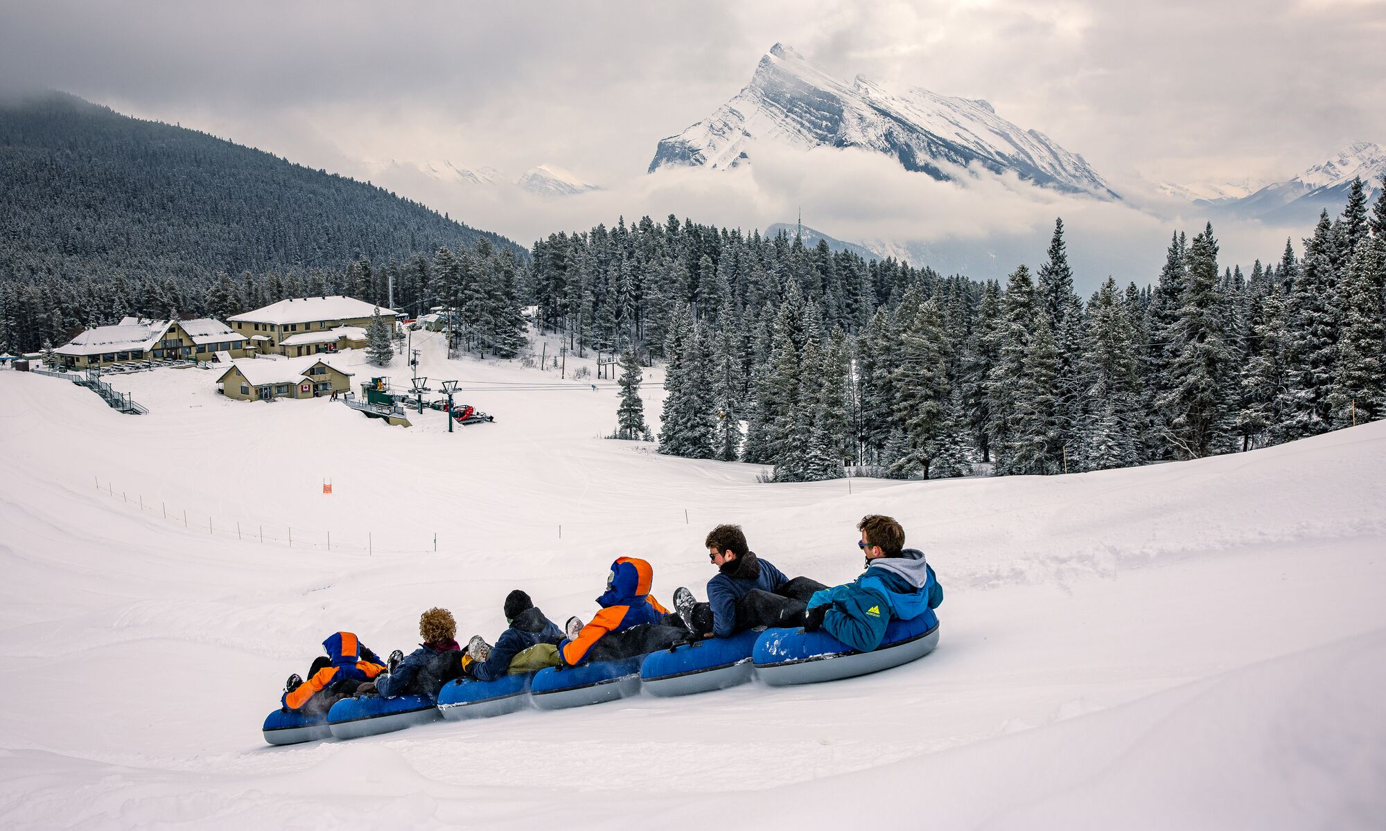 A group of 6 friends tube down a snowy hill together at the Mt. Norquay Ski Resort