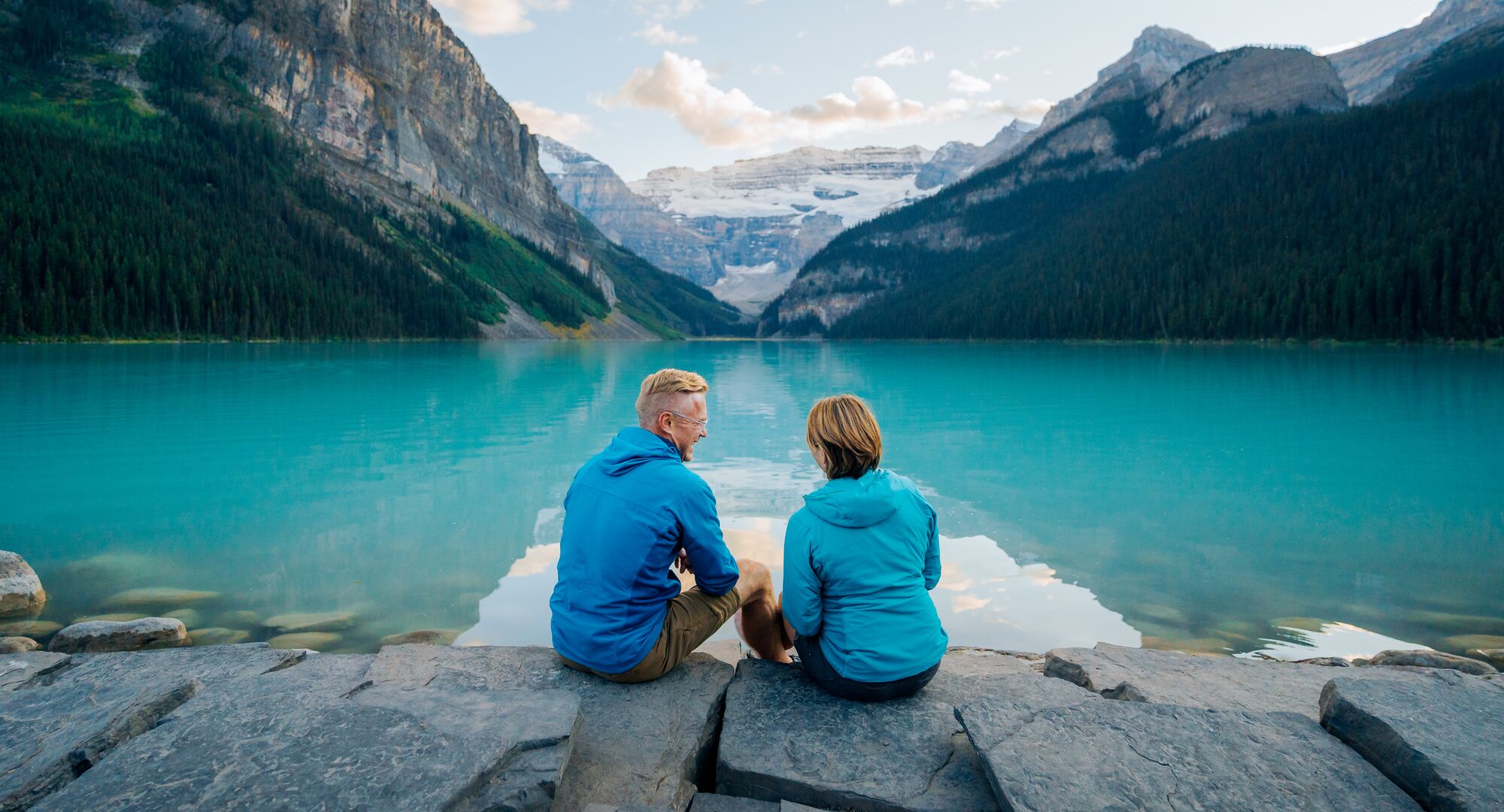 A couple sits on the shore of a turquoise lake staring out at a glacier in the distance while visiting Lake Louise at sunset