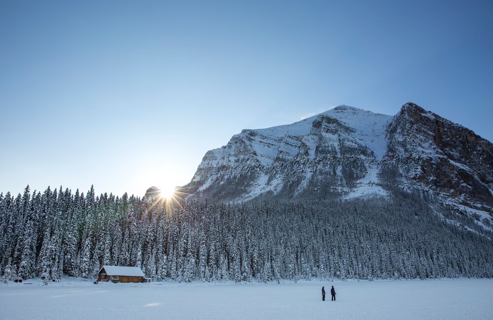 A couple walking by Lake Louise backdropped by mountains and trees on a winter day.