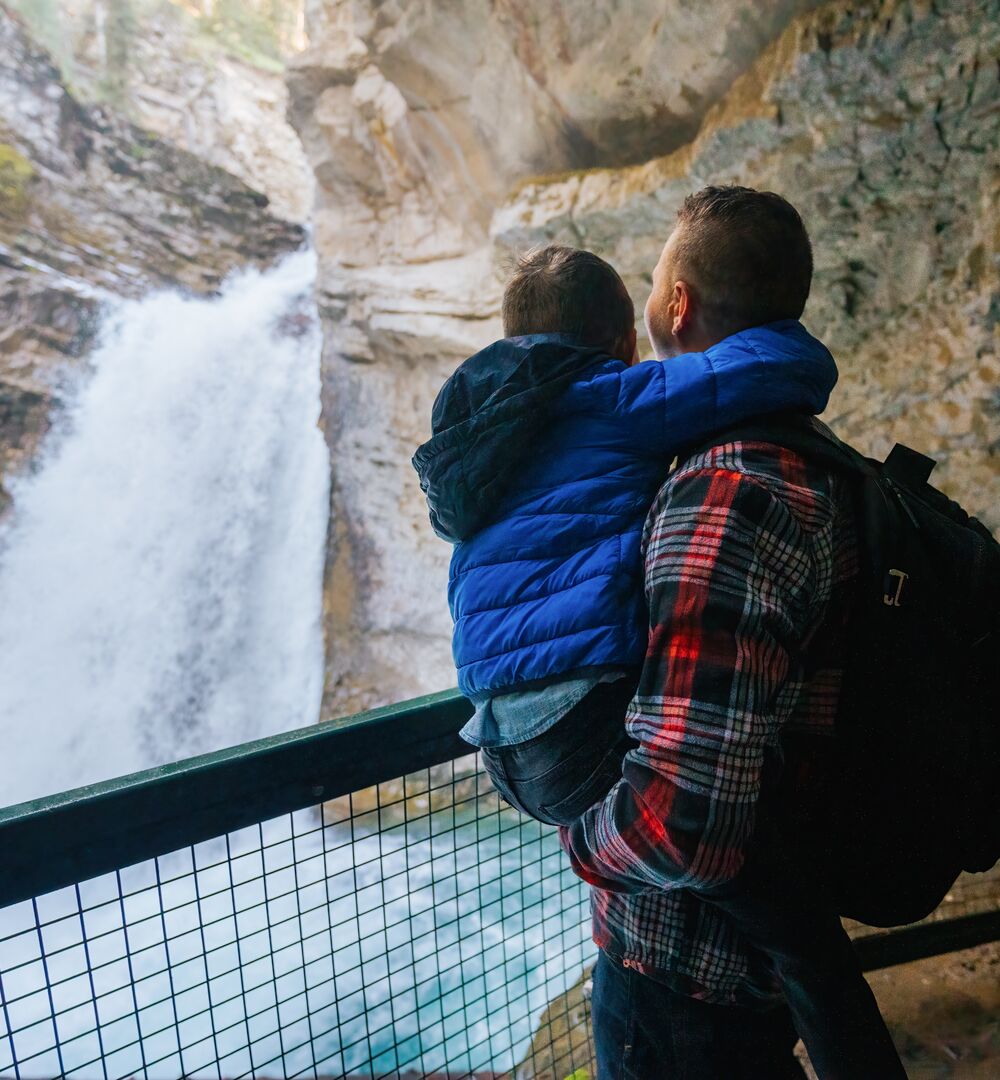 A father wearing a red flannel shirt holds his son in a bright blue jacket as they look at a rushing waterfall from behind the safety of a guardrail at Johnston Canyon in Banff National Park.