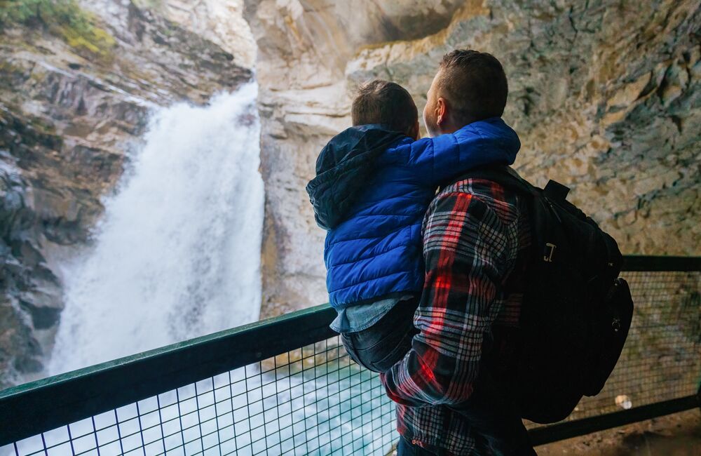 A father wearing a red flannel shirt holds his son in a bright blue jacket as they look at a rushing waterfall from behind the safety of a guardrail at Johnston Canyon in Banff National Park.