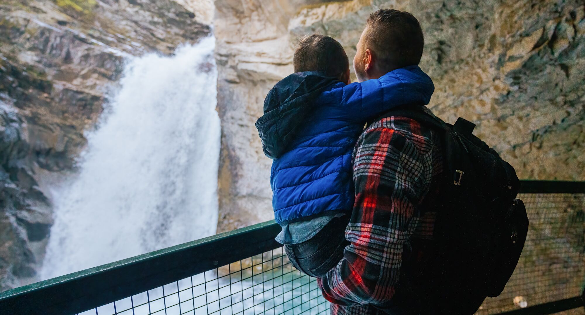 A father wearing a red flannel shirt holds his son in a bright blue jacket as they look at a rushing waterfall from behind the safety of a guardrail at Johnston Canyon in Banff National Park.