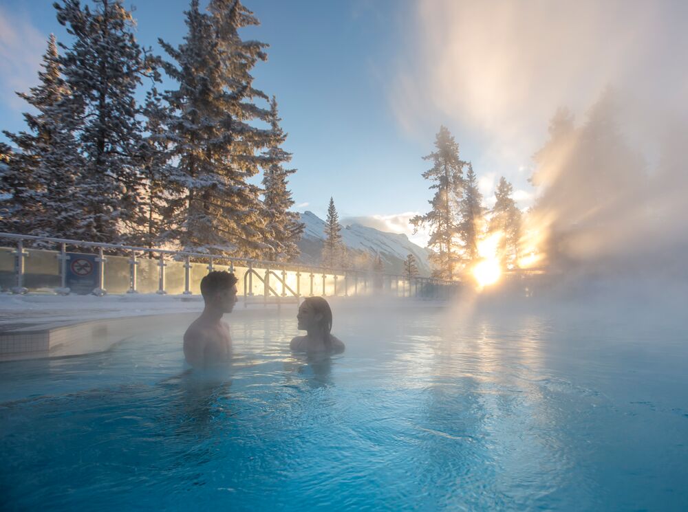 A couple sitting in an outdoor hot pool with snow covered trees in the background in Banff National Park