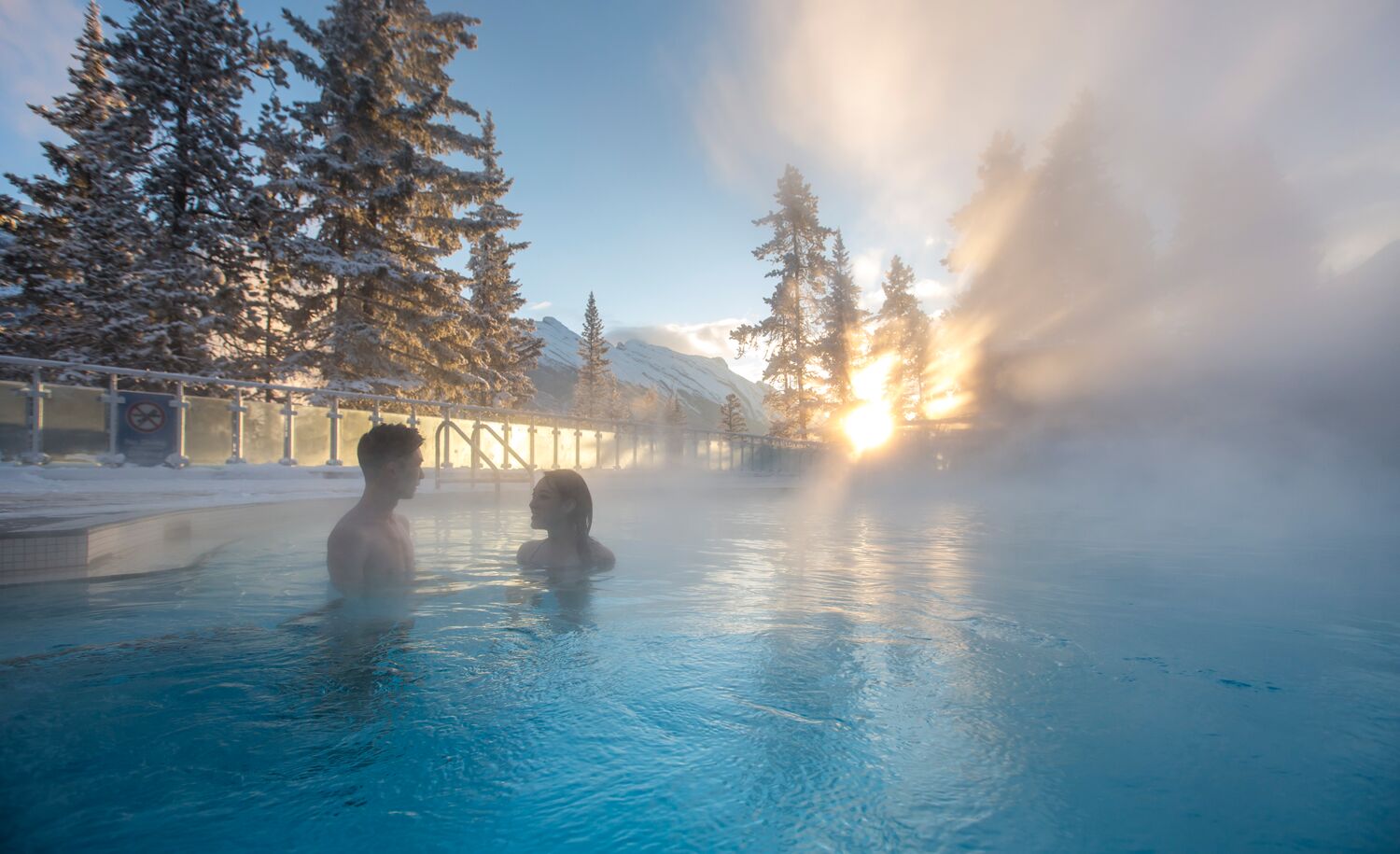 A couple sitting in an outdoor hot pool with snow covered trees in the background in Banff National Park