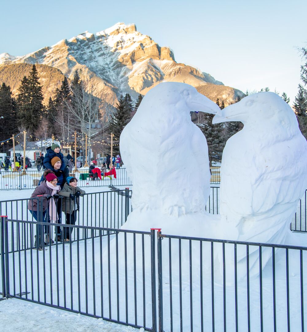 A family looks at a life-sized snow sculpture with Cascade Mountain in the background in Banff National Park.