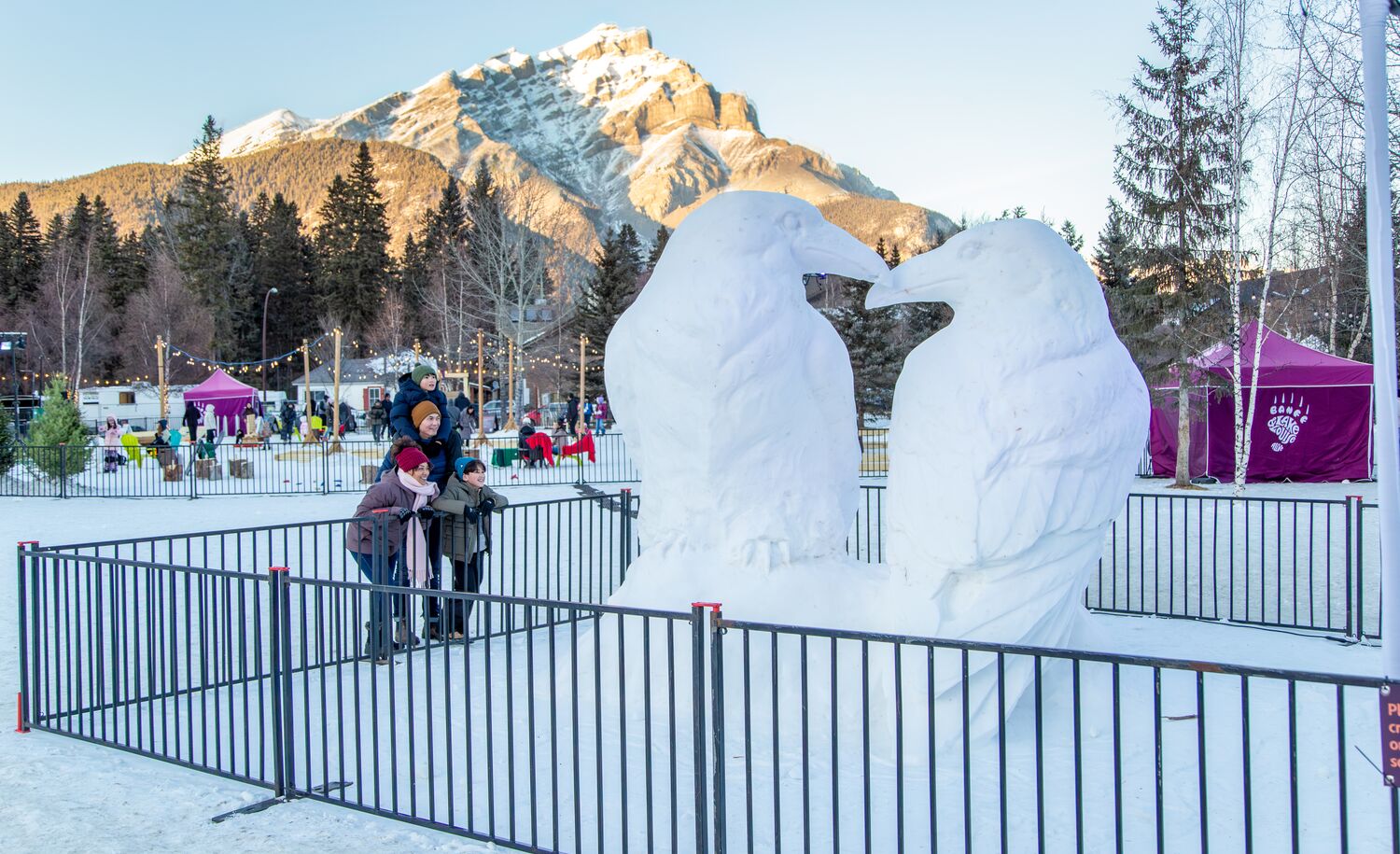 A family looks at a life-sized snow sculpture with Cascade Mountain in the background in Banff National Park.