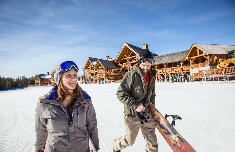 A man and woman walking across a snowy ski resort with the lodge in the background. Both are wearing their goggles as it is a bright sunny day