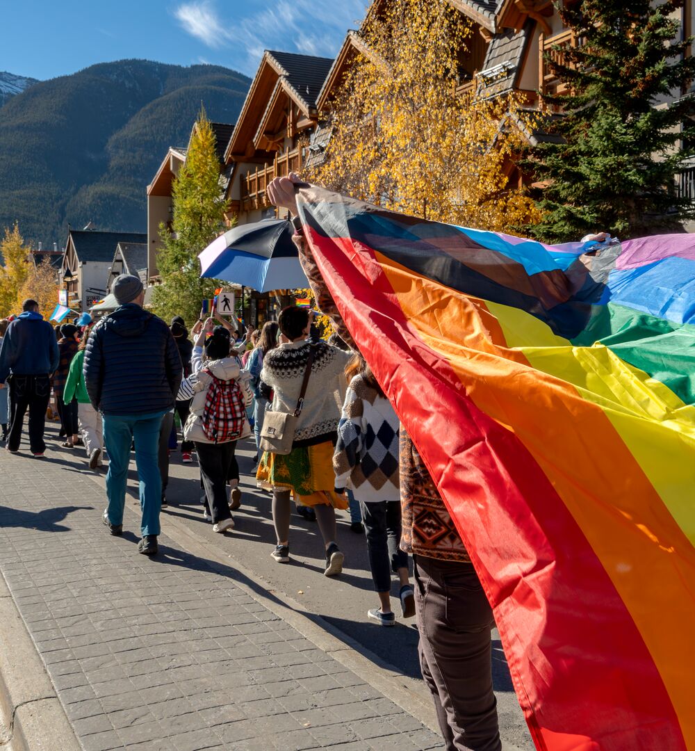 A person swings a pride flag near the camera during the Banff Pride Parade on Banff Ave with mountains in the distance while surrounded by people in Banff in Banff National Park.