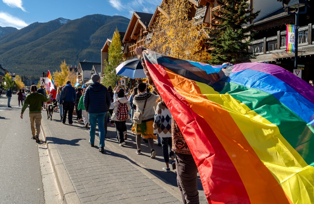 A person swings a pride flag near the camera during the Banff Pride Parade on Banff Ave with mountains in the distance while surrounded by people in Banff in Banff National Park.
