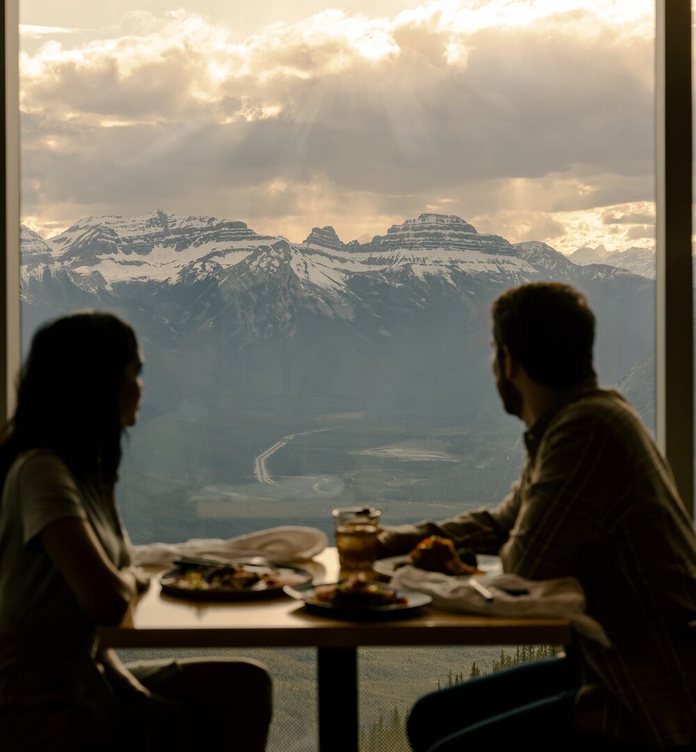 A couple enjoys a meal at the top of the Banff Gondola with views of surrounding mountain peaks in Banff National Park