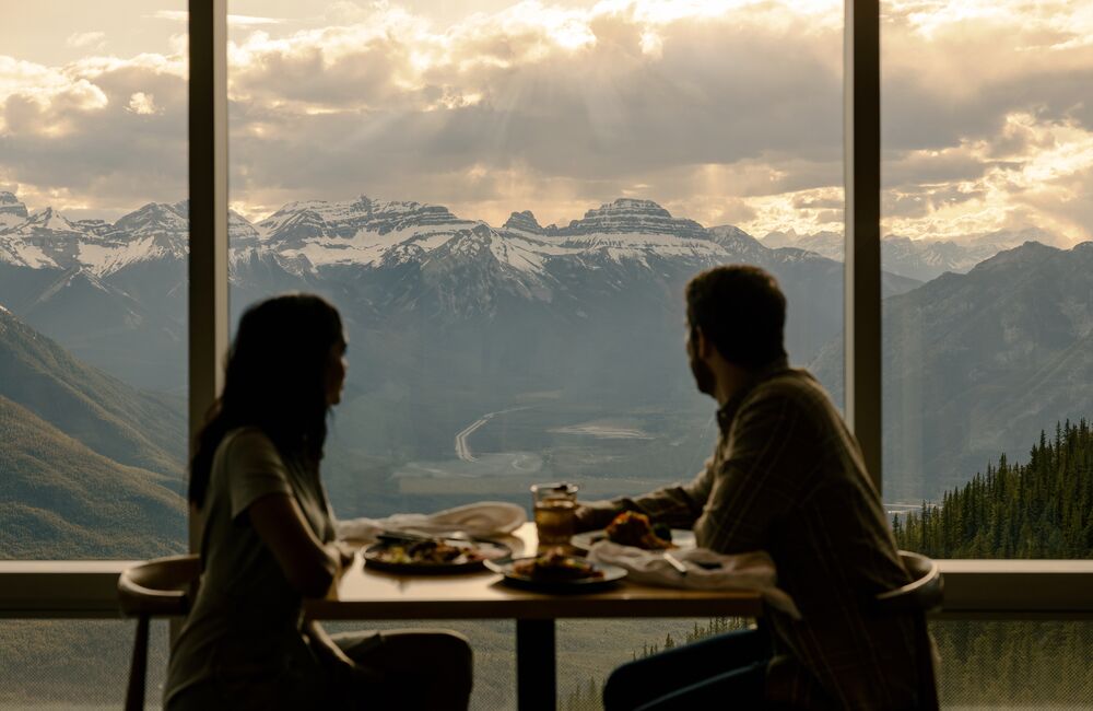 A couple enjoys a meal at the top of the Banff Gondola with views of surrounding mountain peaks in Banff National Park