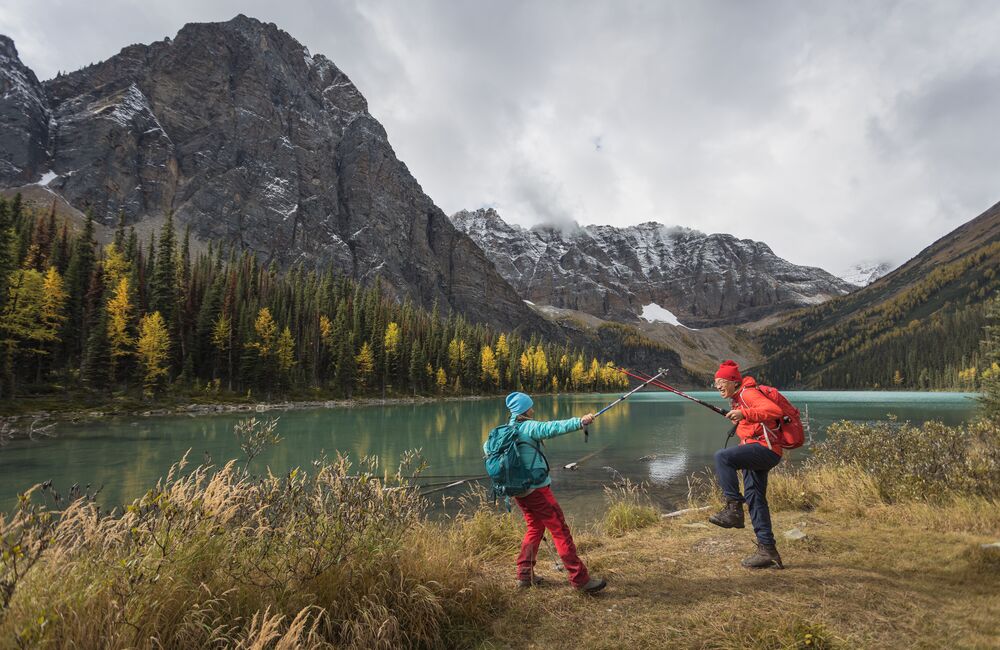 A man and woman hike alongside a lake with yellow larch trees and fresh snow on the mountains surrounding them