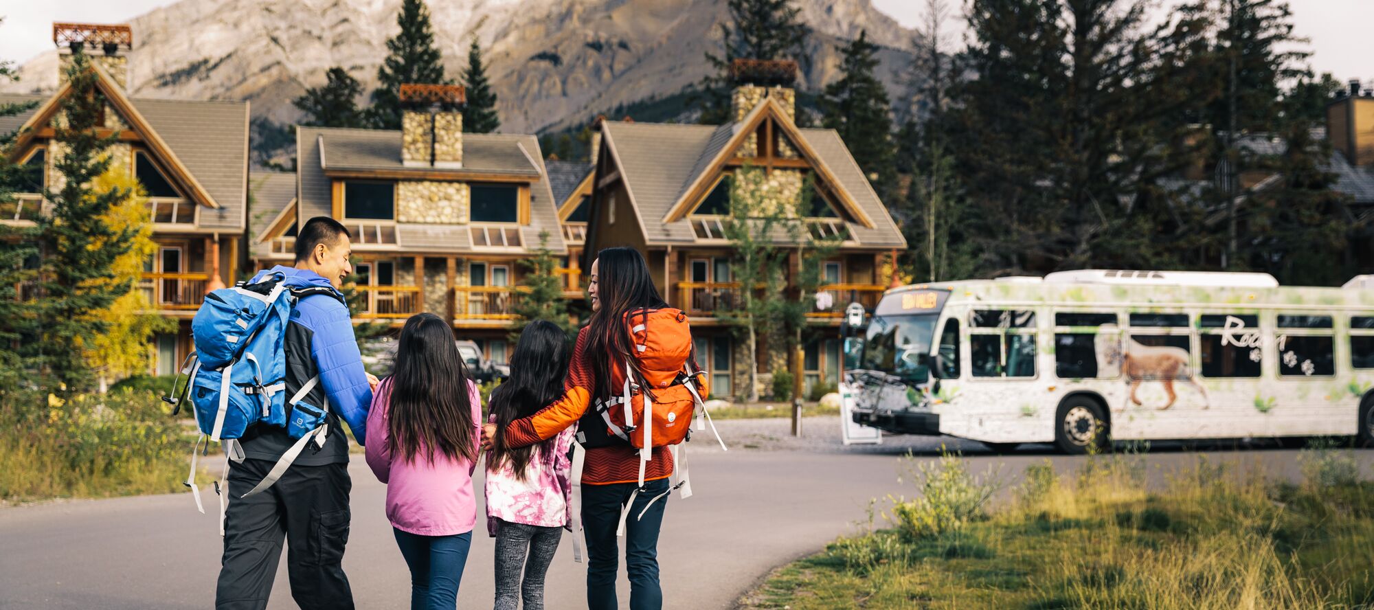 A public Roam transit bus is pulled to the side of the road as three friends take in the view of turquoise Two Jack Lake and Mount Rundle on a sunny summer day in Banff National Park