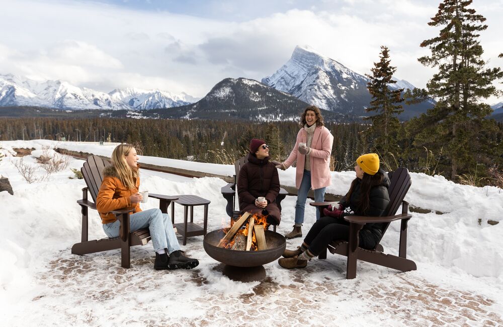 Friends enjoy warm drinks around the camp fire at Juniper Bistro with snowy Mount Rundle behind them in Banff National Park.