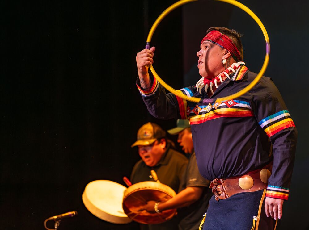 Indigenous man throwing hoops with people drumming in the background wearing Indigenous regalia.