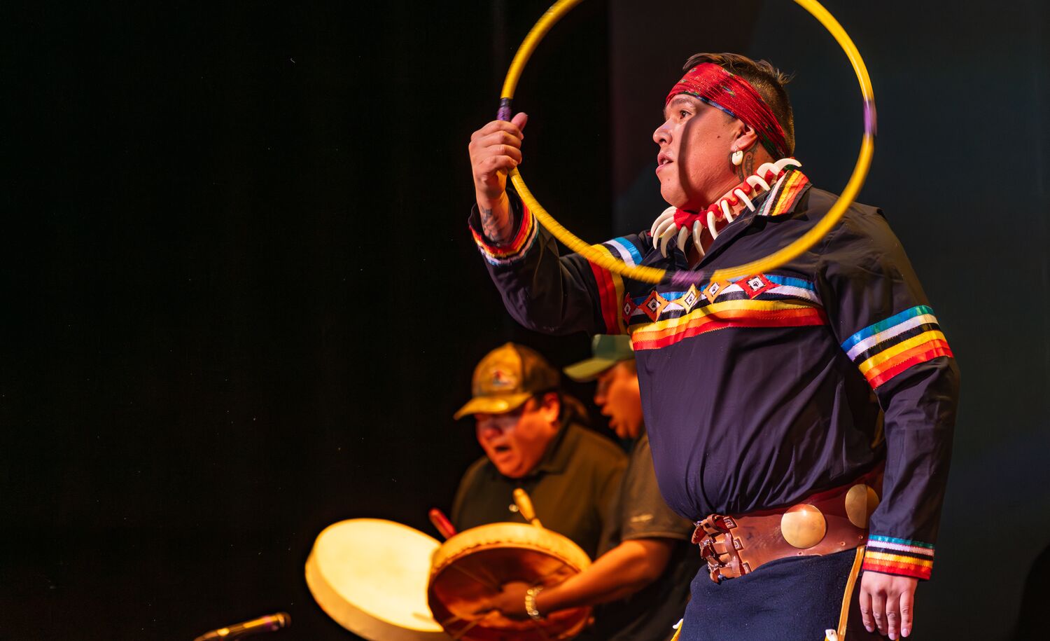Indigenous man throwing hoops with people drumming in the background wearing Indigenous regalia.