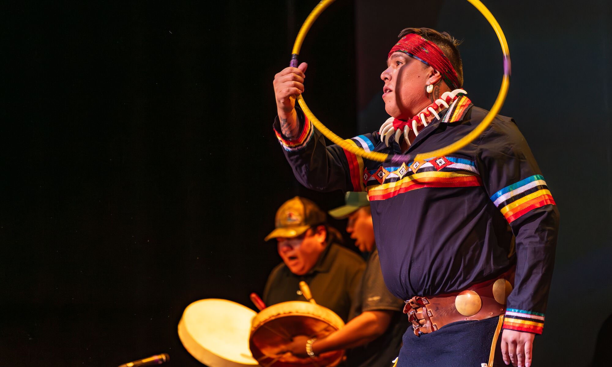 Indigenous man throwing hoops with people drumming in the background wearing Indigenous regalia.