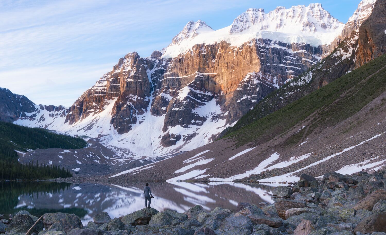 A photo of a clear lake reflecting the trees, snow-capped mountains, and rock formations surrounding it