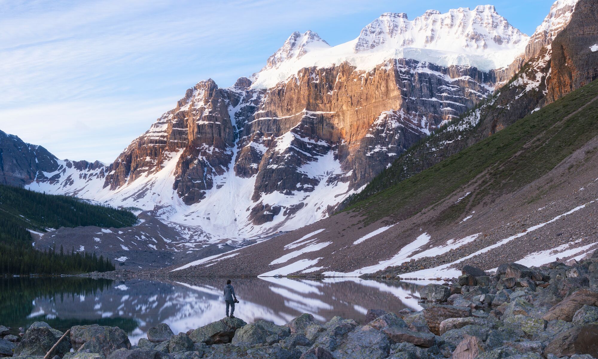 A photo of a clear lake reflecting the trees, snow-capped mountains, and rock formations surrounding it