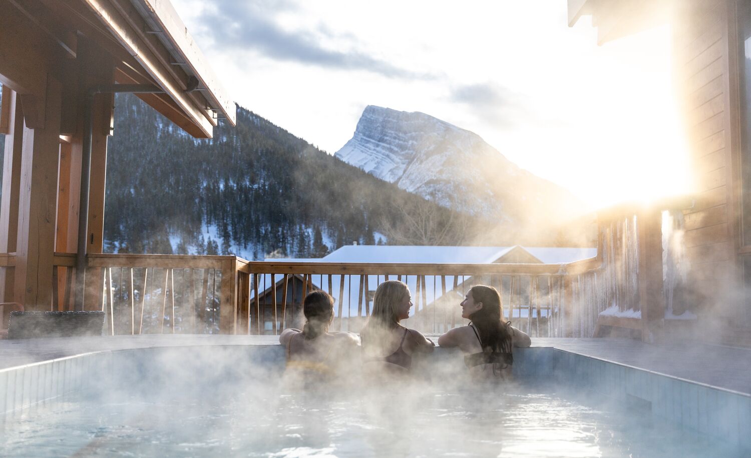 Three women hang out in the houtdoor hot tub at the Moose Hotel in Banff National Park with Mt. Rundle in the background.