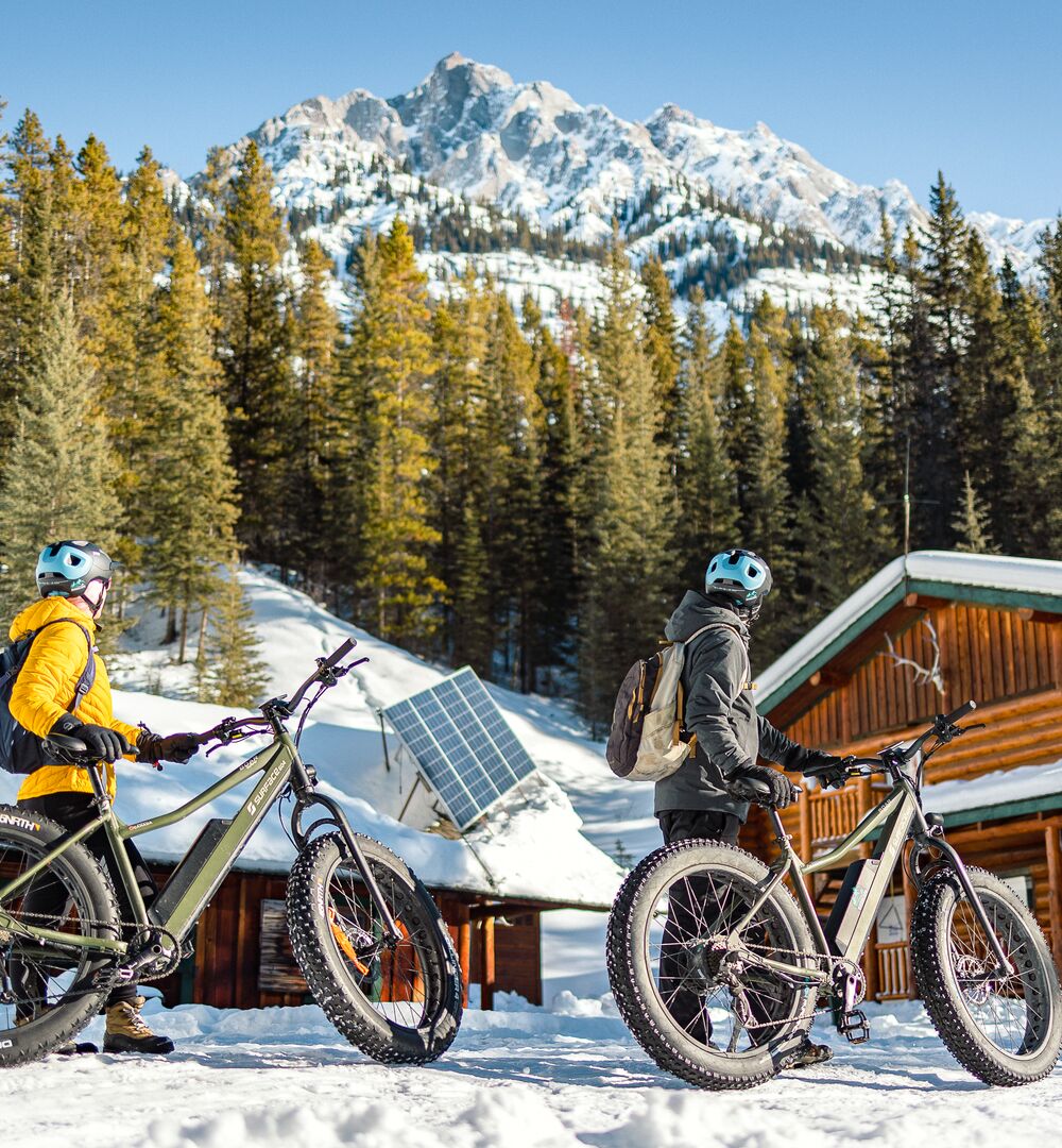 Two people stand with their fat bikes at Sundance Lodge in front of a snowy mountain in Banff National Park.