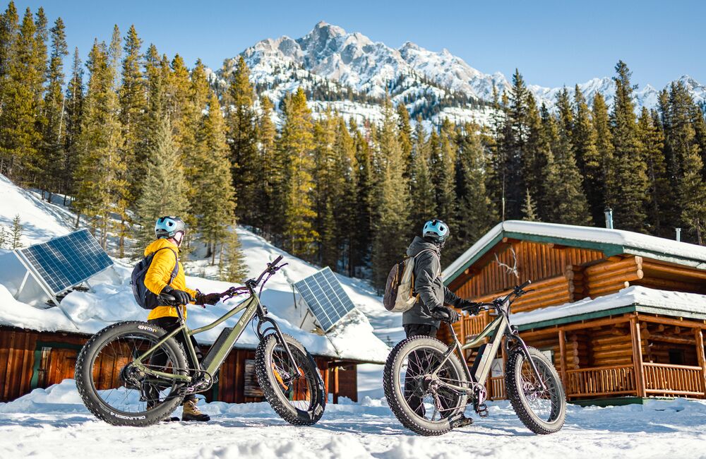 Two people stand with their fat bikes at Sundance Lodge in front of a snowy mountain in Banff National Park.