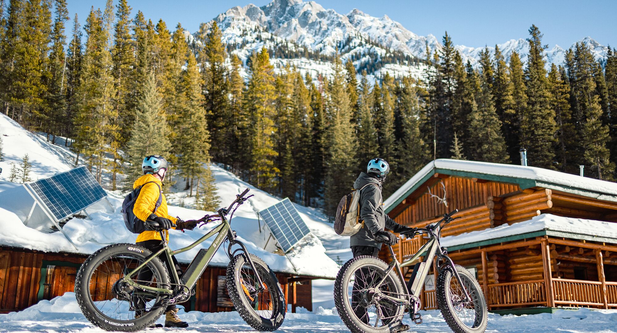 Two people stand with their fat bikes at Sundance Lodge in front of a snowy mountain in Banff National Park.