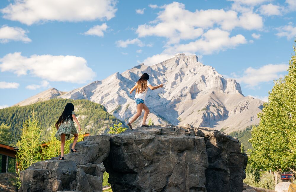 Two young girls run up a rock at the Banff Central Park Playground in the summer with Cascade Mountain in the background in Banff National Park.