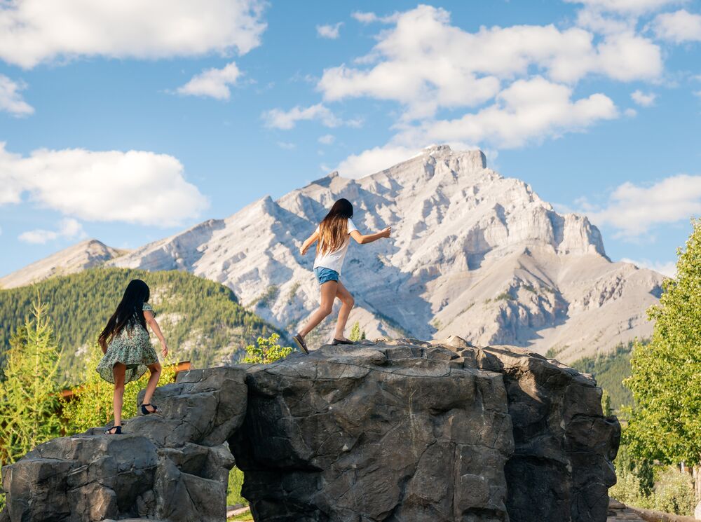 Two young girls run up a rock at the Banff Central Park Playground in the summer with Cascade Mountain in the background in Banff National Park.