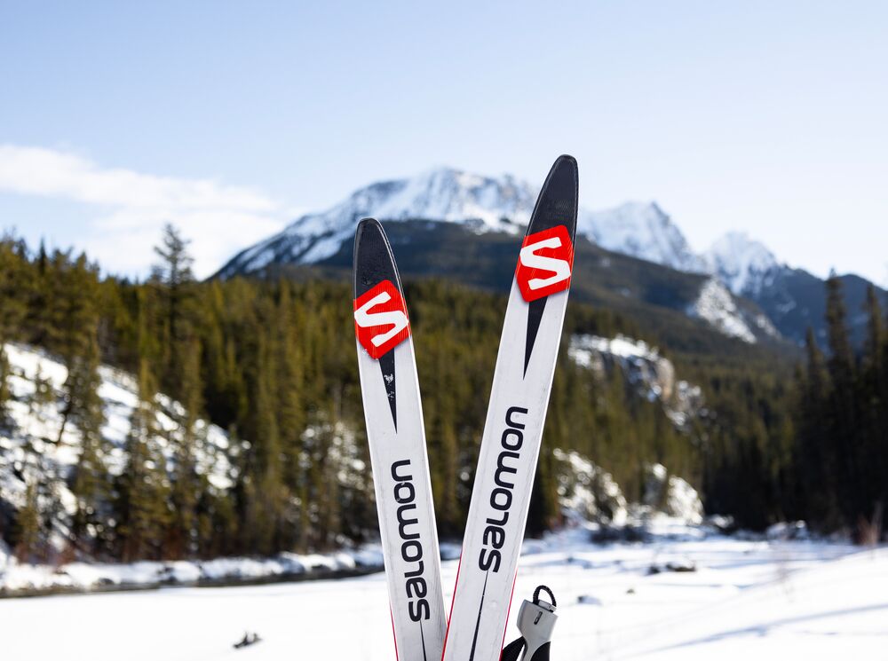 A pair of cross country skis sticking upwards in the snow with a mountain out of focus behind them. 