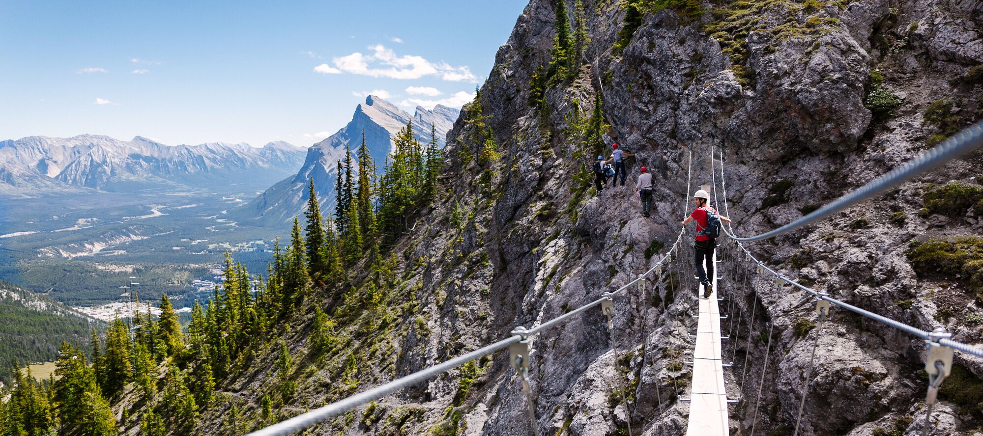 A climber crosses a suspension bridge towards a large mountain while overlooking the valley below at the Via Ferrata
